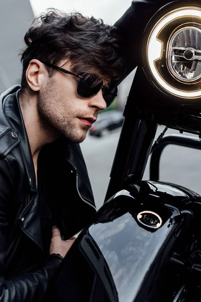 Schöner junger Mann sitzt neben schwarzem Motorrad und lehnt sich kopfüber an Lenker — Stockfoto