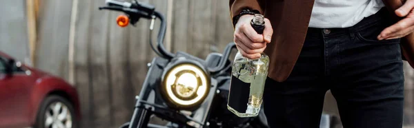 Panoramic shot of man in jacket walking off motorcycle and holding bottle of alcohol — Stock Photo