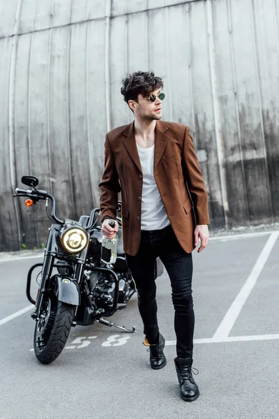 Повний вигляд молодого чоловіка в сонцезахисних окулярах, що стоїть біля мотоцикла і тримає пляшку з алкоголем — стокове фото