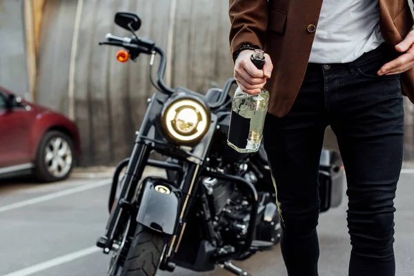 Vista recortada del hombre con chaqueta caminando fuera de la motocicleta y sosteniendo la botella de alcohol - foto de stock