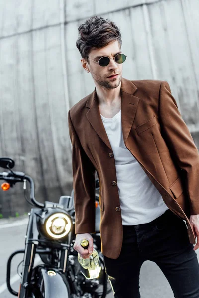 Селективное внимание привлекательного мужчины в солнцезащитных очках и коричневой куртке, идущего с бутылкой алкоголя возле мотоцикла — стоковое фото