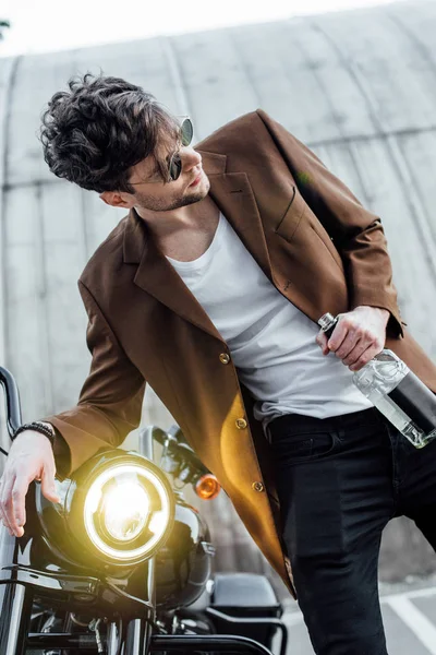 Handsome young man leaning on handlebars of motorcycle with light lamp while holding bottle with alcohol — Stock Photo