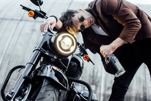 Joven cabeza apoyada en el manillar de la motocicleta con lámpara de luz mientras sostiene la botella con alcohol - foto de stock