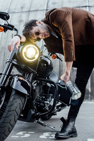 Молодий чоловік спирається головою на кермо мотоцикла з яскравою лампою, тримаючи пляшку з алкоголем — стокове фото