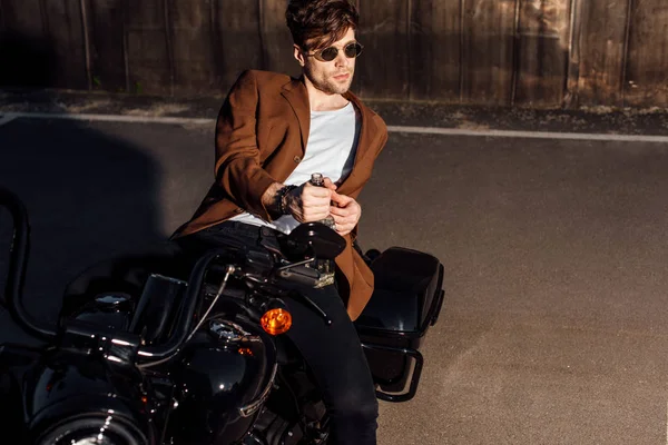 Jovem bonito em casaco marrom deitado na motocicleta e segurando garrafa de álcool — Fotografia de Stock