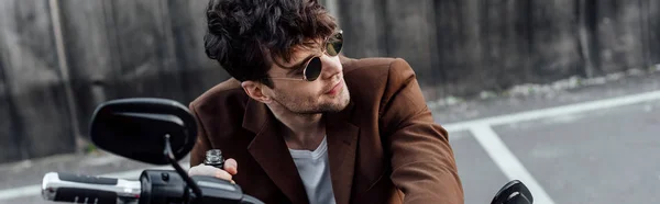 Panoramic shot of man in sunglasses holding bottle, sitting on motorcycle and looking away — Stock Photo