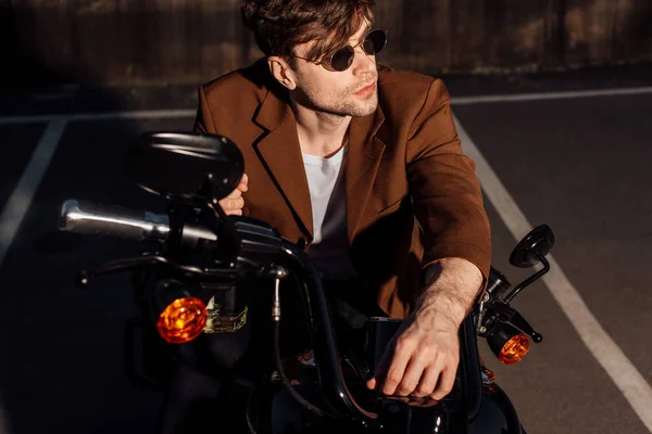 Hombre guapo en gafas de sol sentado en motocicleta y mirando hacia otro lado - foto de stock