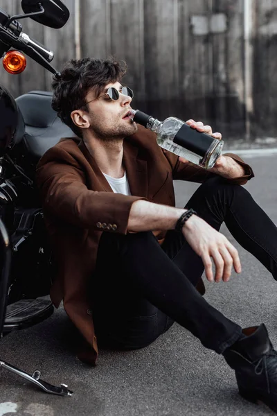 Joven bebiendo alcohol mientras está sentado en el suelo cerca de la motocicleta - foto de stock