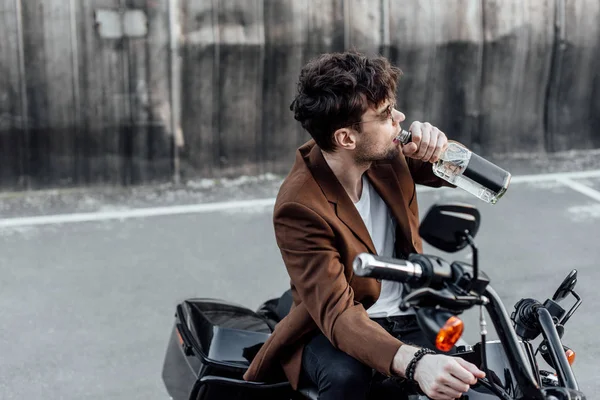 Человек в коричневой куртке пьет алкоголь из бутылки, сидя на мотоцикле — стоковое фото