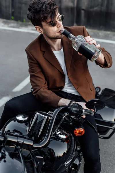 Bel homme en lunettes de soleil et veste brune boire de l'alcool de la bouteille tout en étant assis sur la moto — Photo de stock