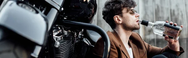 Tiro panorâmico de homem bonito em óculos de sol e casaco marrom beber álcool de garrafa enquanto sentado na motocicleta — Fotografia de Stock