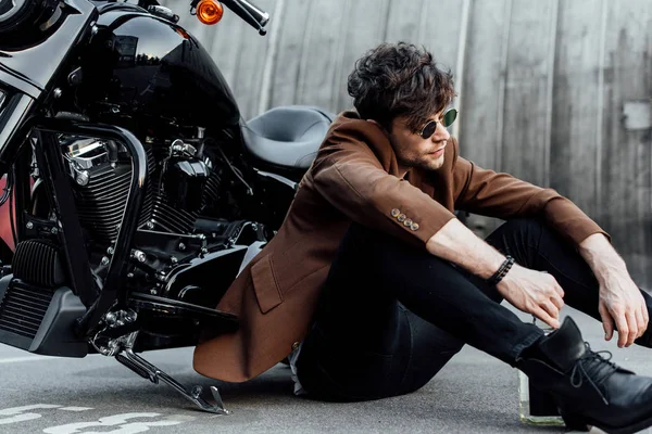 Uomo in elegante giacca marrone seduto a terra vicino alla moto, guardando lontano e tenendo bottiglia di alcol — Foto stock