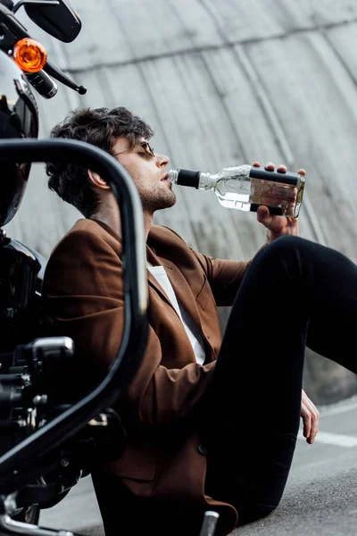 Selective focus of man in sunglasses and brown jacket drinking alcohol from bottle while sitting on motorcycle — Stock Photo