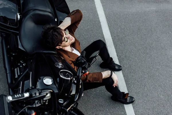 Vista superior del hombre en gafas de sol y traje sentado en el suelo cerca de la motocicleta - foto de stock