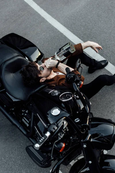 Vue de dessus de l'homme en lunettes de soleil et costume assis sur le sol près de la moto et boire de l'alcool de la bouteille — Photo de stock