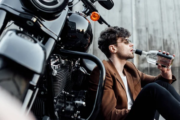 Selektiver Fokus des Mannes, der Alkohol trinkt, während er sich auf das Motorrad stützt und auf dem Boden sitzt — Stockfoto