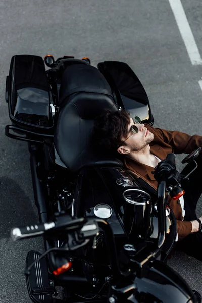 Young man in glasses and brown jacket resting while sitting on ground and leaninghead on motorcyce — Stock Photo