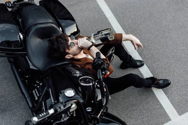 Vue de dessus du jeune motard assis sur le sol, penché sur la moto et buvant de l'alcool — Photo de stock