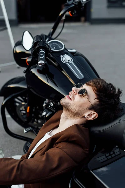 Handsome motorcyclist in brown jacket resting while leaning head on motorcycle — Stock Photo