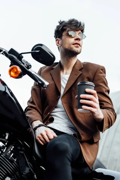 Низкий угол обзора стильного мужчины в куртке и солнцезащитных очках, держащего кофе, чтобы пойти, сидя на мотоцикле — стоковое фото
