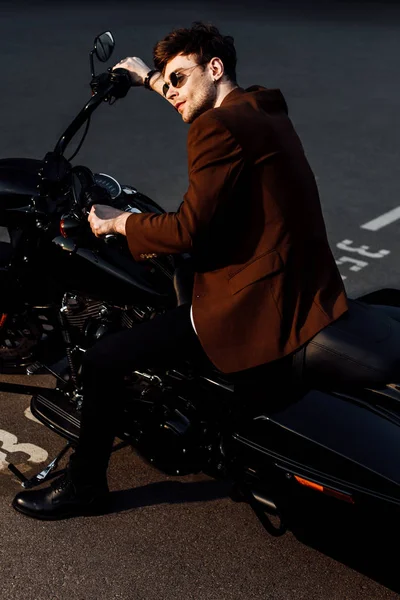 Мотоциклист в коричневой куртке и солнцезащитных очках сидит на мотоцикле и держит руль — стоковое фото