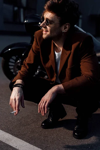 Young man in sunglasses and jacket sitting squatting near motorcycle and smoking cigarette — Stock Photo