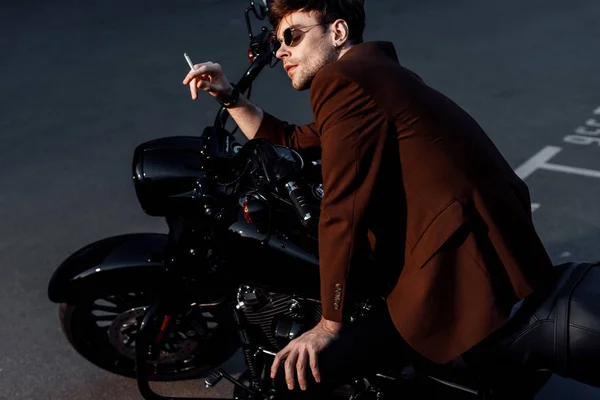 Young man in brown jacket sitting on black motorcycle and smoking cigarette — Stock Photo