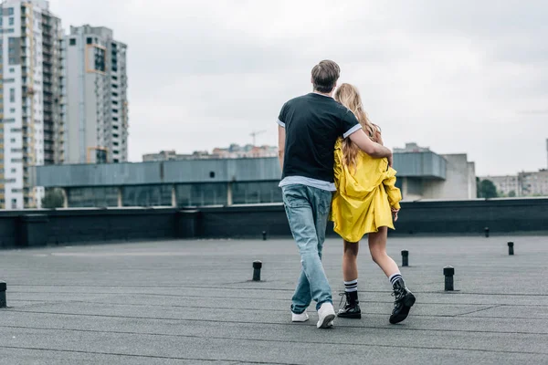 Visão traseira da mulher em casaco amarelo e homem em jeans e t-shirt abraçando no telhado — Fotografia de Stock