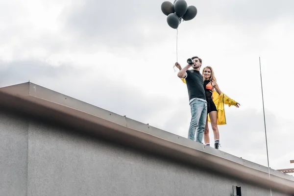 Atractiva mujer sosteniendo globos negros y guapo hombre bebiendo champán - foto de stock