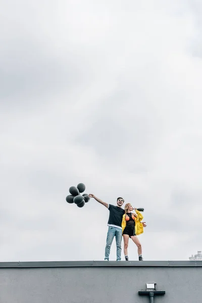 Atractiva mujer bebiendo champán y guapo hombre sosteniendo globos negros - foto de stock