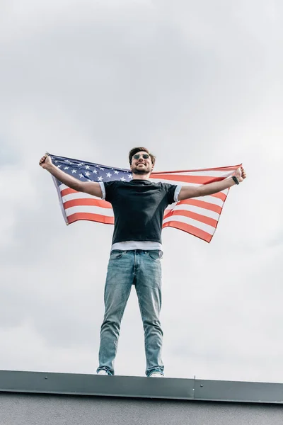 Красивый мужчина в очках держит американский флаг и улыбается на крыше — стоковое фото