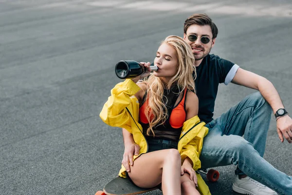 Attractive woman drinking champagne and handsome man in glasses sitting on skateboard — Stock Photo