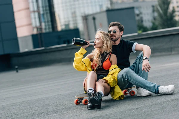Attrayant femme boire du champagne et bel homme dans des lunettes assis sur skateboard — Photo de stock