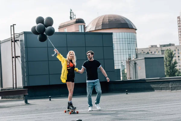 Attractive woman with balloons skateboarding and man in glasses holding her hand — Stock Photo