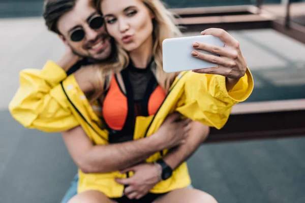 Hermosa mujer en chaqueta amarilla tomando selfie con hombre guapo - foto de stock