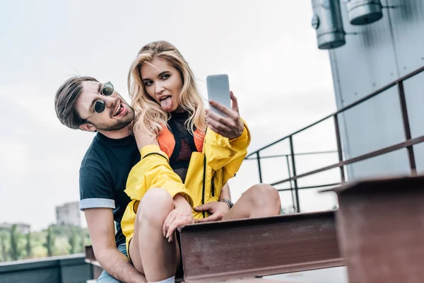 Atractiva y rubia mujer sobresaliendo lengua y tomando selfie con hombre guapo - foto de stock