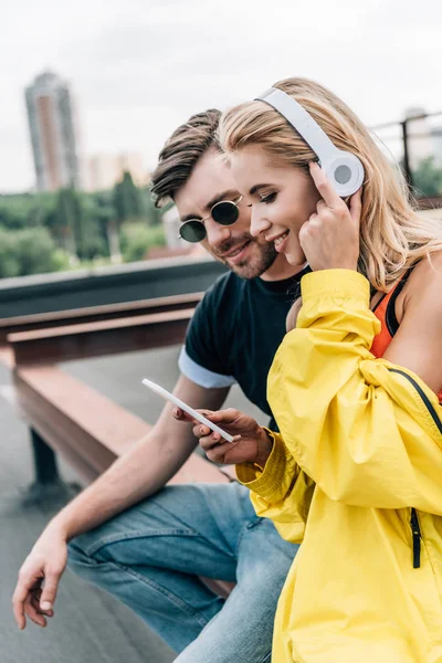 Привлекательная блондинка с наушниками, использующая смартфон, и мужчина, смотрящий на него — стоковое фото