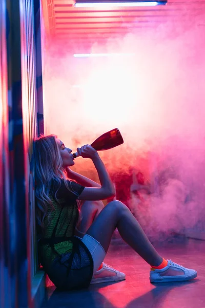 Вид збоку приваблива і блондинка сидить на підлозі і п'є шампанське — стокове фото