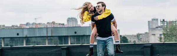 Panoramic shot of attractive and blonde woman and handsome man hugging and playing on roof — Stock Photo