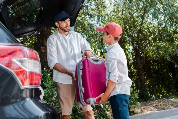 Избирательный фокус счастливого отца, стоящего рядом с сыном с багажом возле багажника автомобиля — стоковое фото