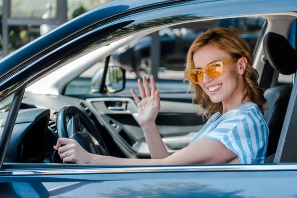 Весела жінка в сонцезахисних окулярах махає рукою під час водіння автомобіля — стокове фото