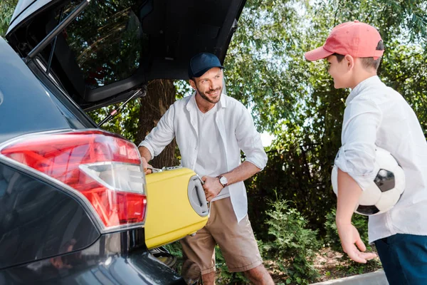 Foco seletivo do homem feliz em cap colocando saco de viagem no porta-malas do carro perto do filho — Fotografia de Stock