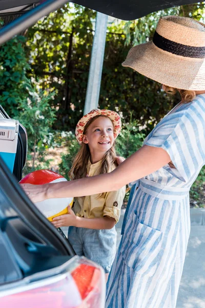 Селективное внимание веселого ребенка, смотрящего на мать в соломенной шляпе возле машины — стоковое фото