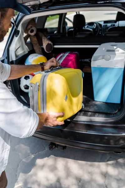 Cropped view of man putting yellow luggage near pink travel bag in car trunk — Stock Photo