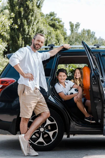 Счастливый отец стоит с рукой на бедре рядом с детьми в машине — стоковое фото