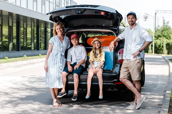 Hombre feliz con la mano en la cadera cerca del coche, esposa alegre e hijos - foto de stock