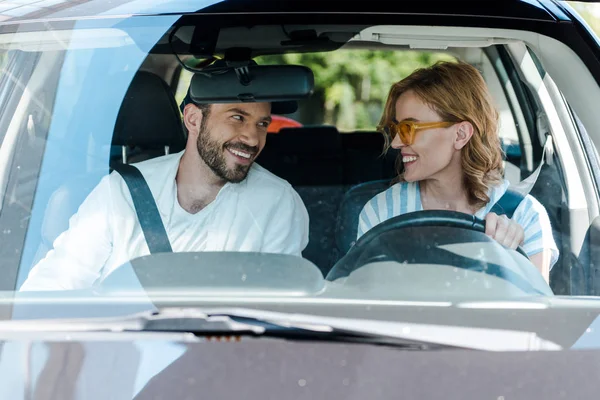 Selective focus of happy woman in yellow sunglasses looking at bearded man in cap while sitting in car — Stock Photo