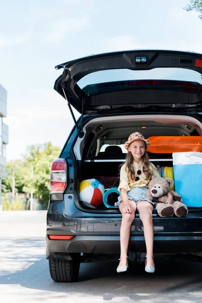Criança feliz segurando ursinho de pelúcia e sentado no porta-malas do carro perto do edifício — Fotografia de Stock