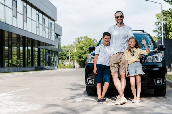 Padre feliz en gafas de sol de pie cerca del coche con los niños - foto de stock