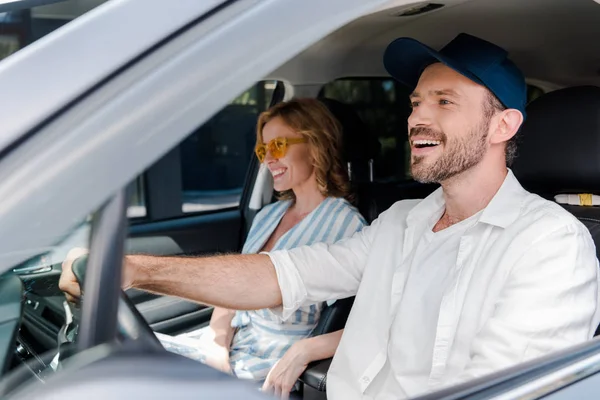 Messa a fuoco selettiva di felice uomo guida auto vicino donna attraente in occhiali da sole — Foto stock